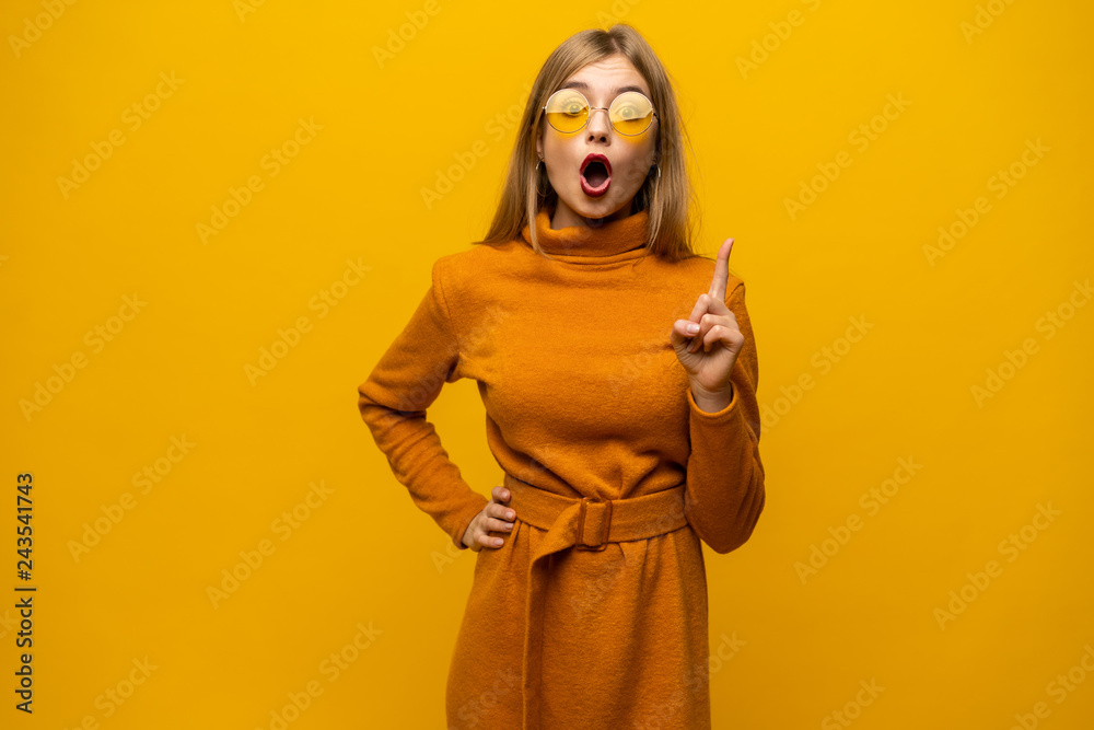 Cheerful lovely young woman standing and pointing away over yellow background.