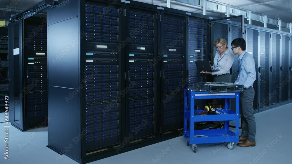 In the Modern Data Center: Engineer and IT Specialist Work with Server Racks, on a Pushcart Equipment for Installing New Hardware. Specialists Doing Maintenance and Diagnostics of the Database. In the Modern Data Center: Engineer and IT Specialist Work with Server Racks, on a Pushcart Equipment for Installing New Hardware. Specialists Doing Maintenance and Diagnostics of the Database.