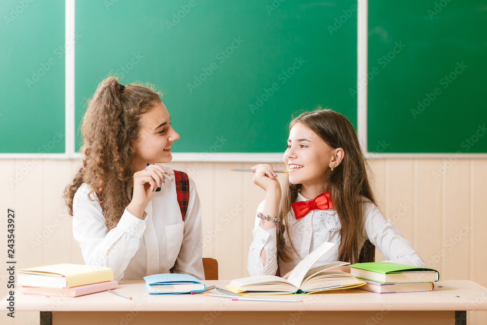 two brunette schoolgirls sit in the classroom at her desk with books on the background of the blackboard two brunette schoolgirls sit in the classroom at her desk with books on the background of the blackboard