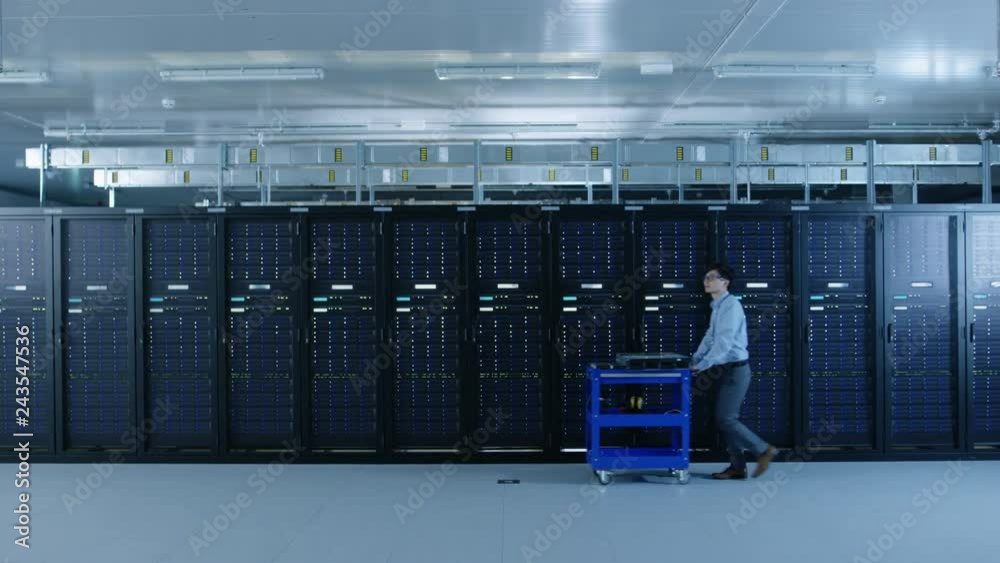 In the Modern Data Center: IT Technician Working with Server Racks, Pushes Cart Between Rows of Server Racks. On a Pushcart New Hardware for System Update. Engineer Doing Maintenance and Diagnostics. In the Modern Data Center: IT Technician Working with Server Racks, Pushes Cart Between Rows of Server Racks. On a Pushcart New Hardware for System Update. Engineer Doing Maintenance and Diagnostics.