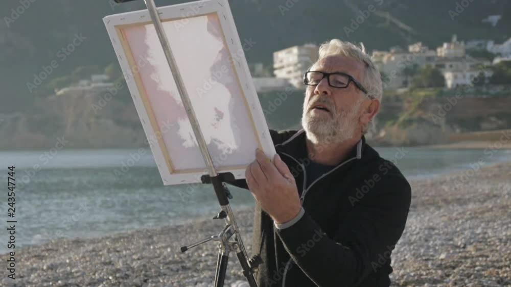 Senior man paints a picture on the beach. Elderly male artist in glass applying paint on white canvas on metal easel at sunset sea beach.