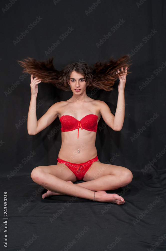 Fashion model posing in bra and red briefs on a uniform black background.  Sitting plays with