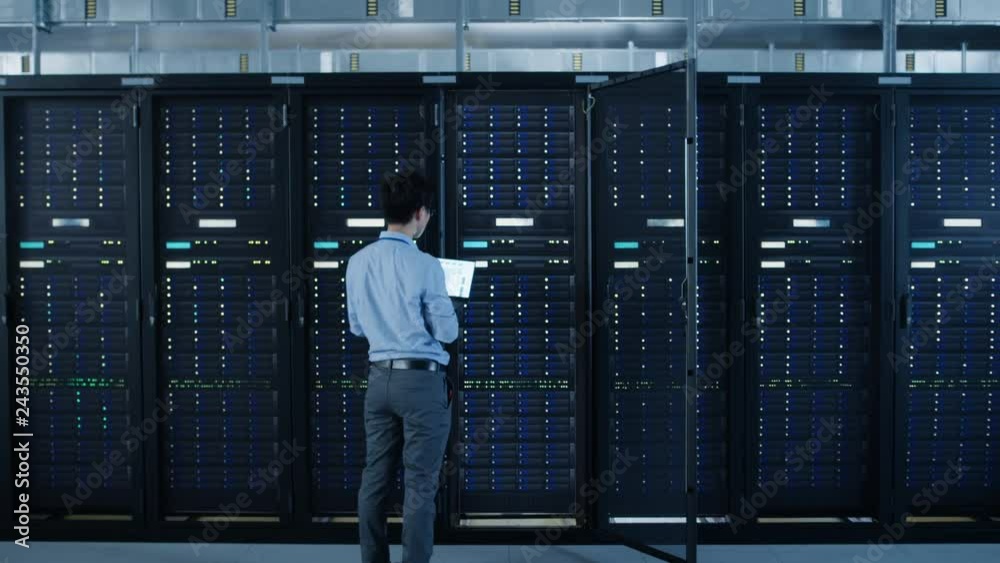  In the Modern Data Center: IT Engineer Standing Beside Open Server Rack Cabinets, Does Wireless Maintenance and Diagnostics Procedure with a Laptop. Zoom Out Camera Shot.