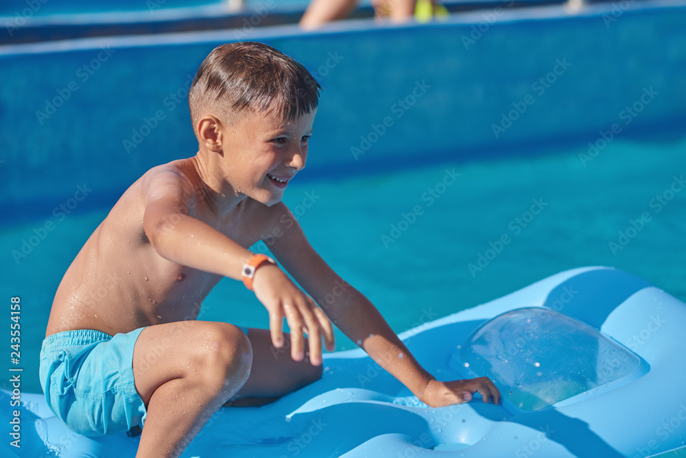 Happy European boy having fun in swimming pool at resort. He is balancing on inflatable mattress.