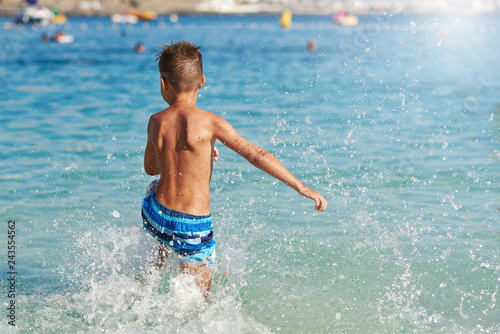 Active European boy in a striped swimming shorts is running towards the sea. He is having fun spending his holidays on the seashore.
