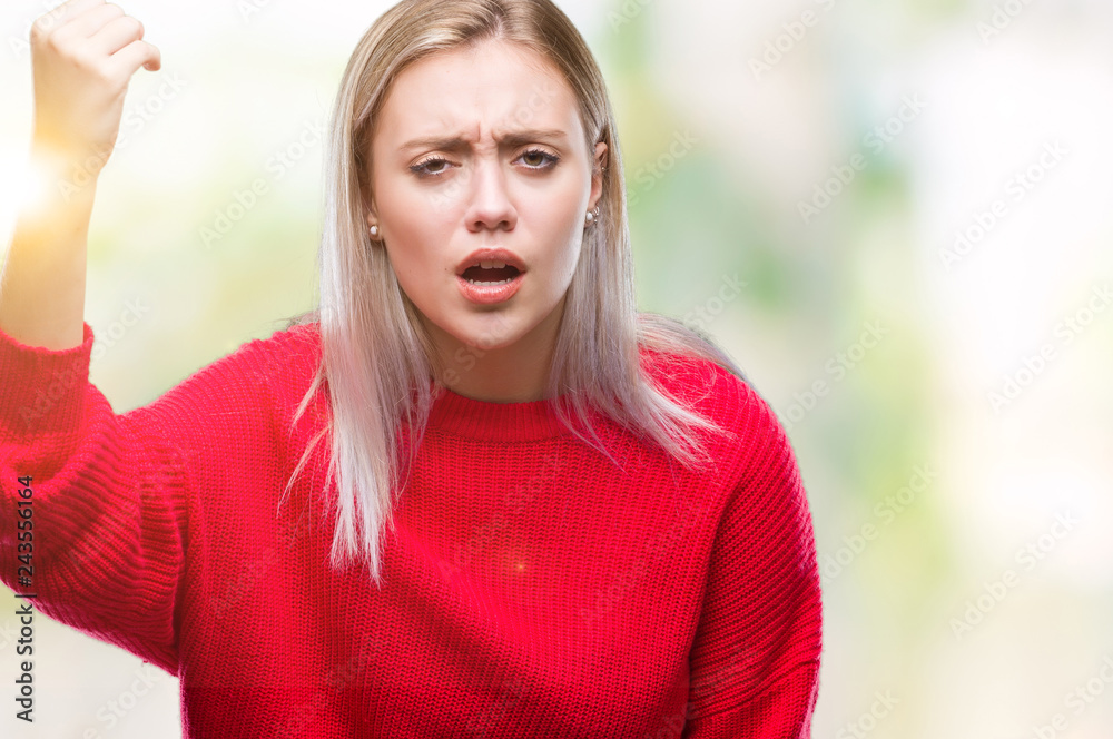 Young blonde woman wearing winter sweater over isolated background angry and mad raising fist frustrated and furious while shouting with anger. Rage and aggressive concept.
