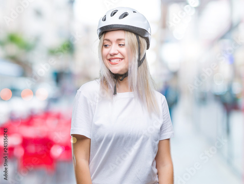 Young blonde woman wearing cyclist security helmet over isolated background looking away to side with smile on face, natural expression. Laughing confident.