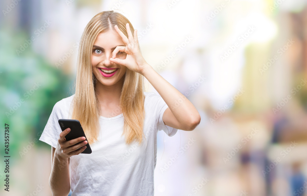 Young beautiful blonde woman using smartphone over isolated background with happy face smiling doing ok sign with hand on eye looking through fingers