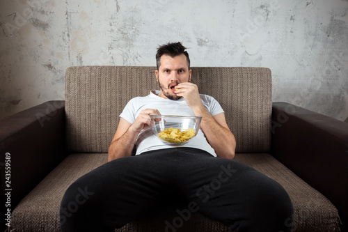 The guy in the shirt is lying on the couch, eating chips and watching a sports channel. The concept of laziness, frustration, procrastination, the person at home. photo