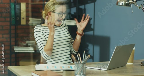 Young pretty Caucasian blond woman in glasses working at the computer in the office and then being very cheerful and happy as seeing some great news or work result. photo