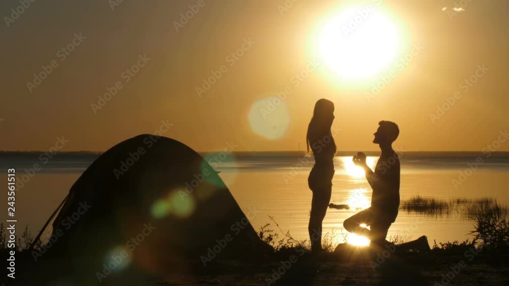 Silhouettes of kneeled man proposing his beloved woman on the beach at sunset. Boyfriend putting engagement ring on happy surprised female's finger. Couple embracing in happiness