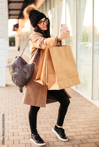 Young trendy woman blogger with shopping bag and smartphone near mall