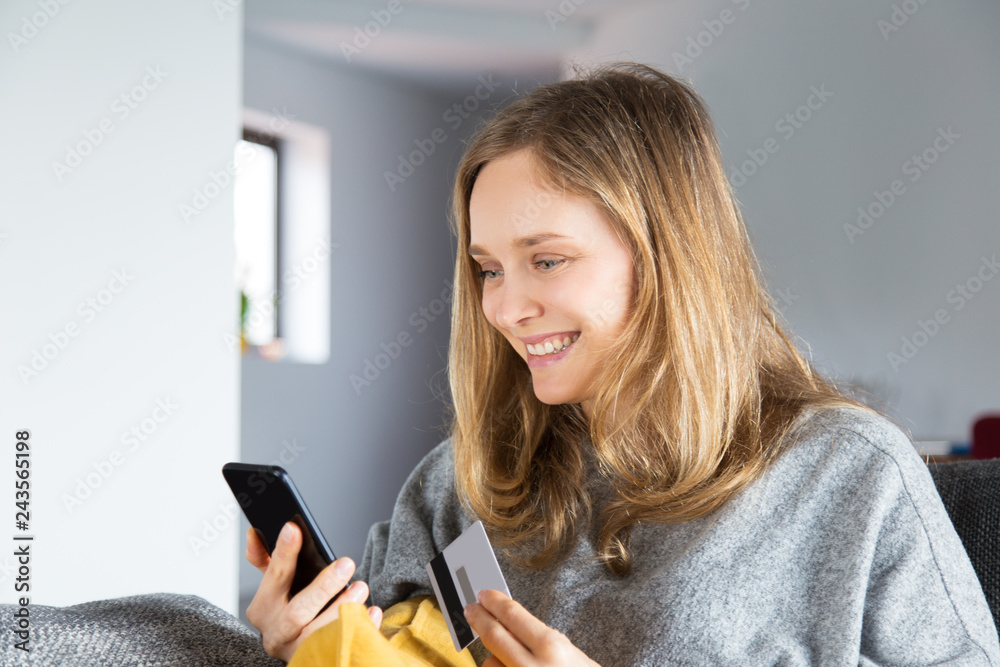 © Mangostar - Happy customer purchasing online and paying by credit card. Cheerful Caucasian girl sitting on couch at home, using phone and holding credit card. Online shopping concept