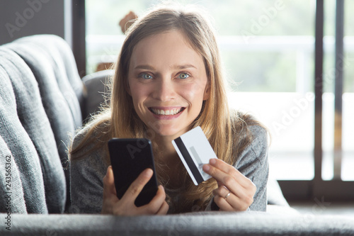 Happy shopaholic paying online by credit card. Cheerful young woman resting on couch at home, holding phone and credit card and smiling at camera. Online payment concept