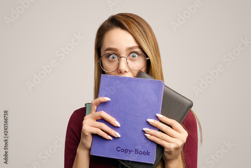 Foto Surprised emotive European woman hides behind books, has eyes popped out, wears round transparent glasses
