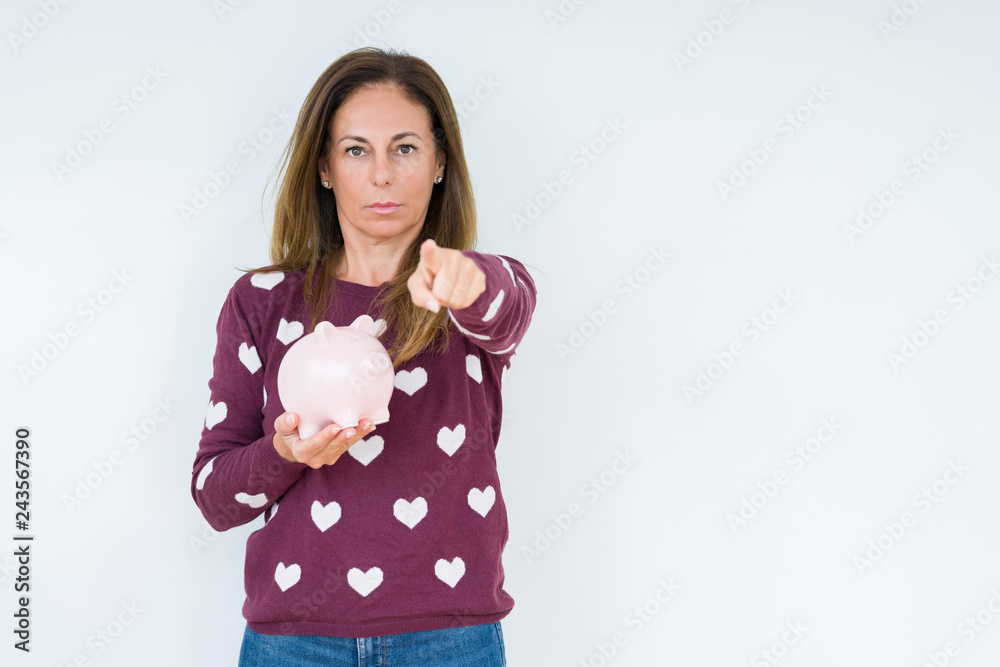 Middle age woman holding piggy bank over isolated background pointing with finger to the camera and to you, hand sign, positive and confident gesture from the front Middle age woman holding piggy bank over isolated background pointing with finger to the camera and to you, hand sign, positive and confident gesture from the front