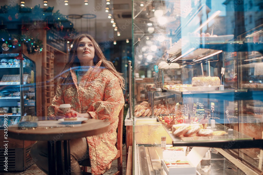 A young woman is sitting behind a window  in a pastry shop cafe with coffee and cake.