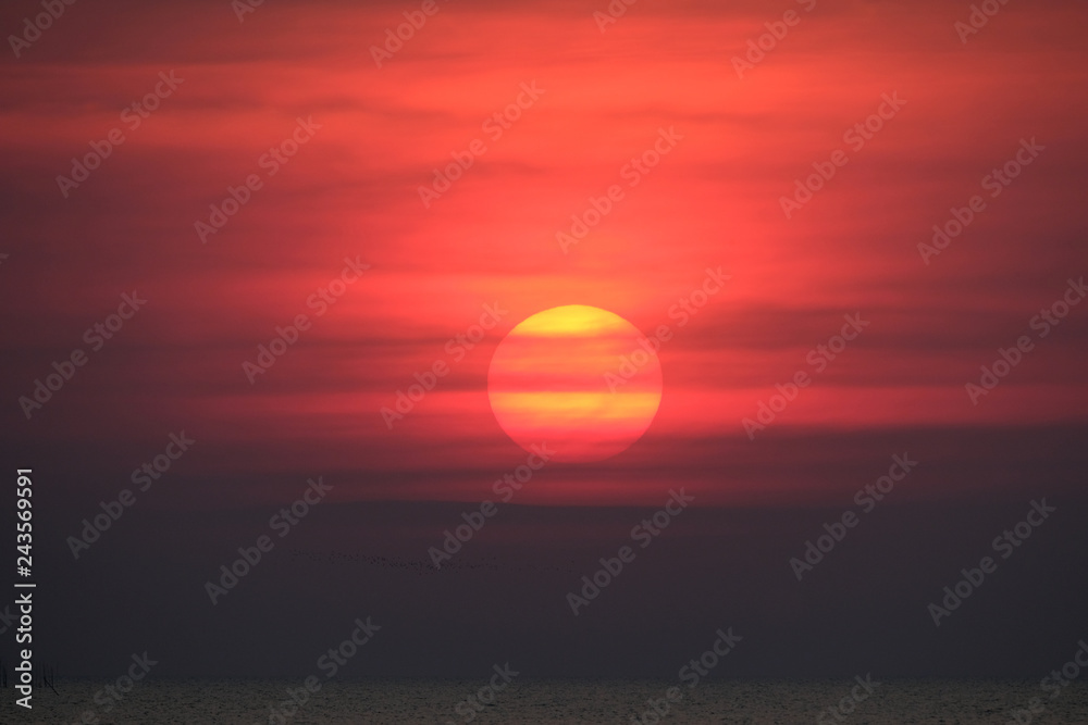 Impressive sunset over the sea with dramatic red fiery sky and dark clouds. Huge glowing orange red sun nearly touch the horizon. Amazing beautiful romantic evening at the beach and perfect vacation. 