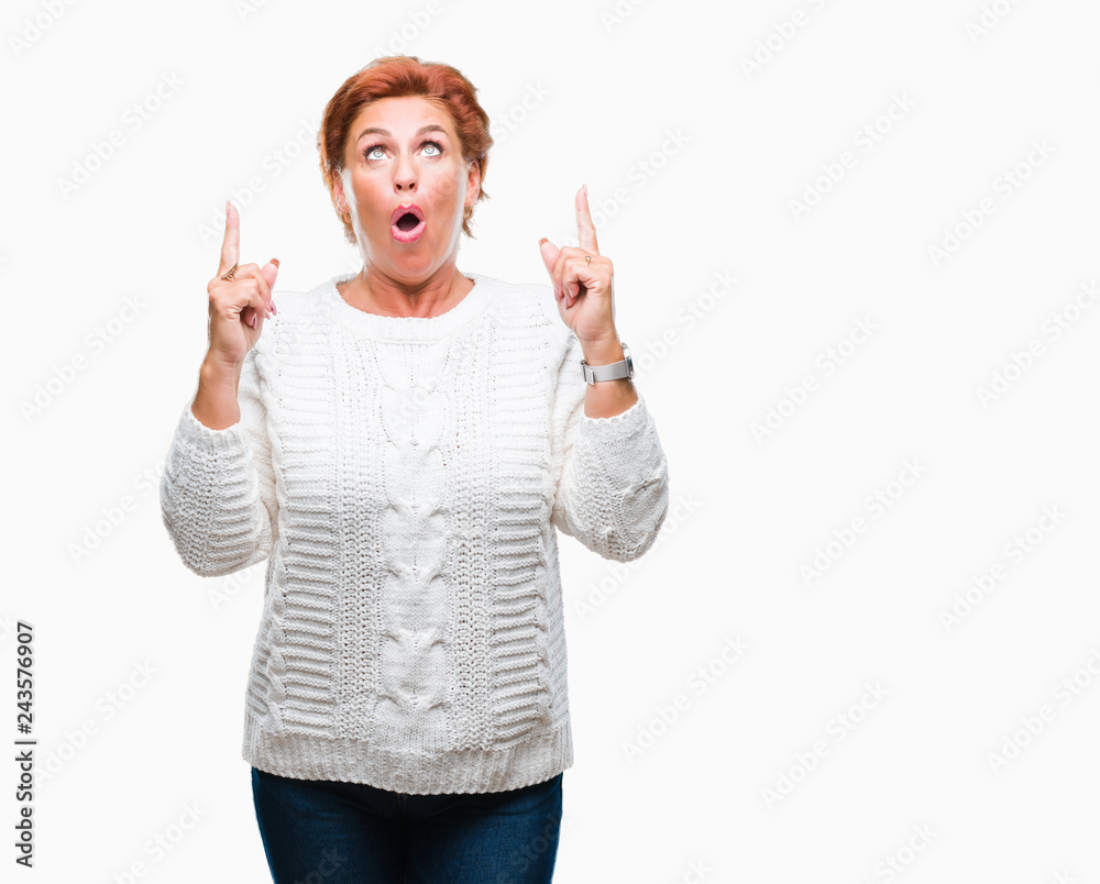 Atrractive senior caucasian redhead woman wearing winter sweater over isolated background amazed and surprised looking up and pointing with fingers and raised arms.