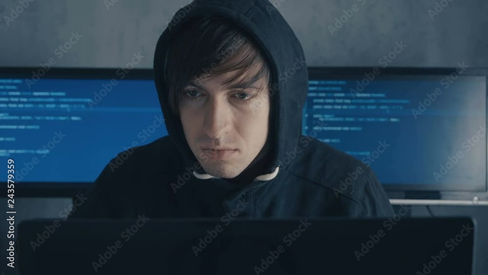 Man in hood sitting at table and coding on laptop. Hacker in hoody working at computer in data center filled monitor screens Man in hood sitting at table and coding on laptop. Hacker in hoody working at computer in data center filled monitor screens