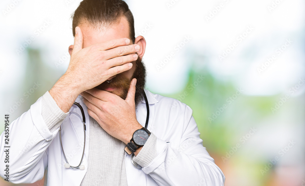 Young blond doctor man with beard wearing medical coat Covering eyes and mouth with hands, surprised and shocked. Hiding emotion Young blond doctor man with beard wearing medical coat Covering eyes and mouth with hands, surprised and shocked. Hiding emotion