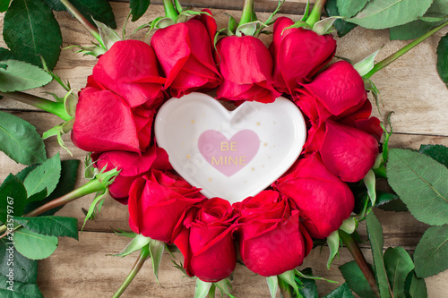 Roses laid out in the shape of a heart on a wooden background, among them a plate in the shape of a heart and with the inscription - Be mine. Festive background for Valentine's Day and March 8th.