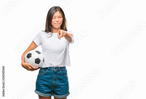 Young asian woman holding football soccer ball over isolated background pointing with finger to the camera and to you, hand sign, positive and confident gesture from the front
