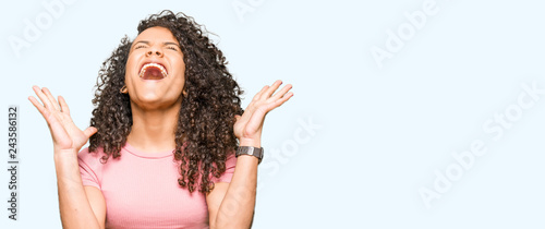 Young beautiful woman with curly hair wearing pink t-shirt celebrating mad and crazy for success with arms raised and closed eyes screaming excited. Winner concept photo