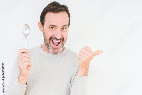 Senior man holding silver spoon over isolated background pointing and showing with thumb up to the side with happy face smiling