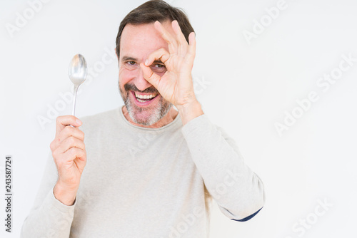 Senior man holding silver spoon over isolated background with happy face smiling doing ok sign with hand on eye looking through fingers