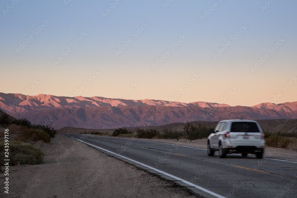 White SUV Vehicle Traveling Down an Empty Desert Road at Sunset or Sunrise with Mountains in the Distance White SUV Vehicle Traveling Down an Empty Desert Road at Sunset or Sunrise with Mountains in the Distance