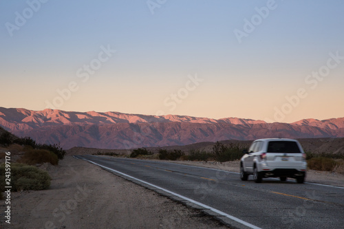 White SUV Vehicle Traveling Down an Empty Desert Road at Sunset or Sunrise with Mountains in the Distance