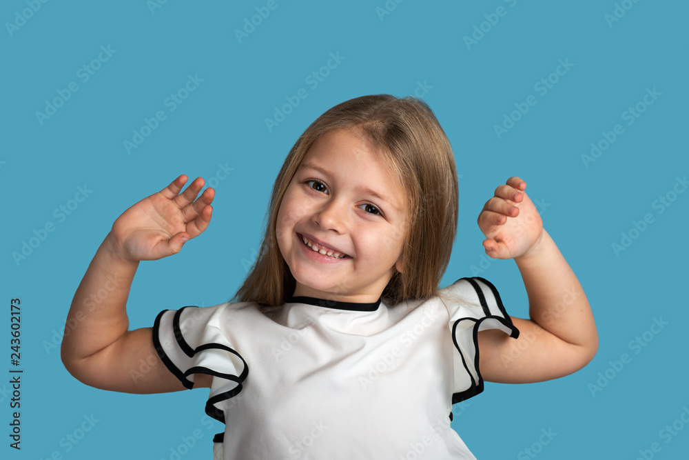 Close up emotional portrait of young blonde  smiling girl wearing white blous with black strips on blue background in studio. She is happy about something having raised her hands up.