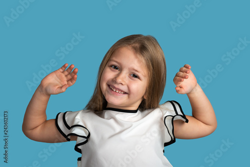 Close up emotional portrait of young blonde smiling girl wearing white blous with black strips on blue background in studio. She is happy about something having raised her hands up.