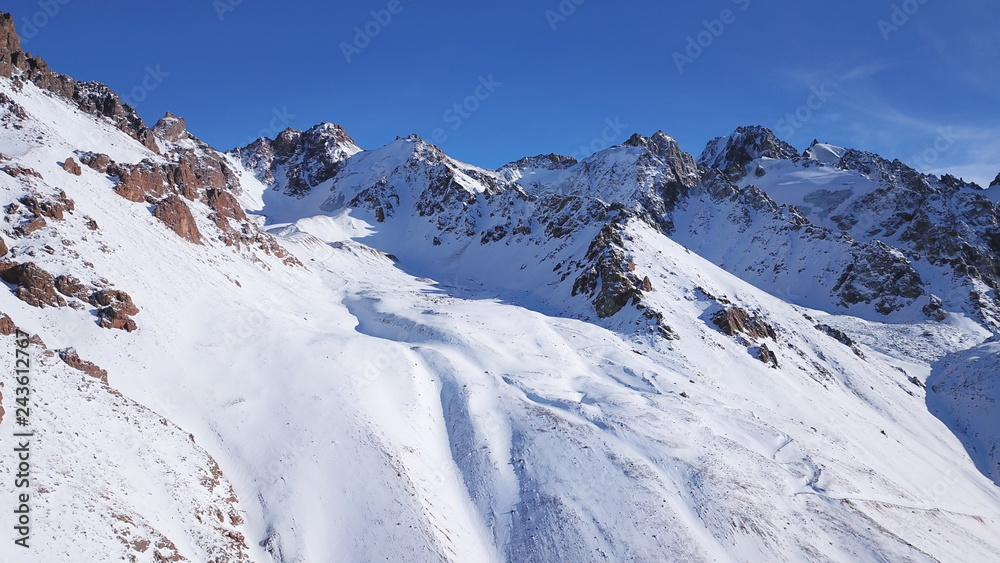 Mountain peaks covered with snow. Blue sky with light white clouds. Through the snow visible rocks and stones. Sharp cliffs and gorge. Snowy mountains shooting with drone. The shade from the sun.