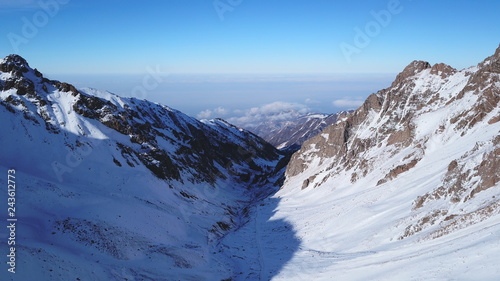 View from the snowy gorge in the mountains to the clouds and the city smog. Coniferous trees and firs are visible. Snowy mountains and grey ground. Gradient blue sky and dark clouds. Drone. © SergeyPanikhin