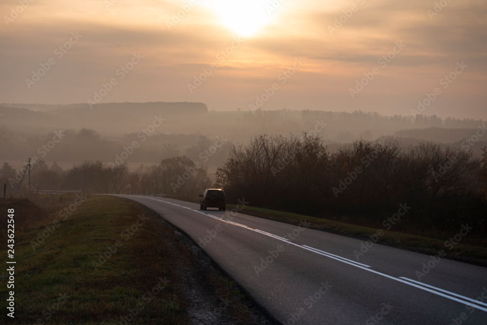 Car traveling on the asphalt road in a rural autumn landscape at sunset
