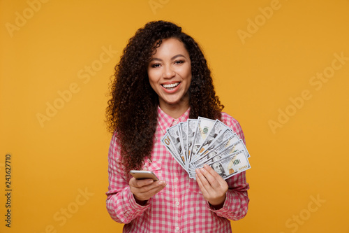 Portrait of an excited young african isolated over yellow background. Looking camera showing display of mobile phone holding money