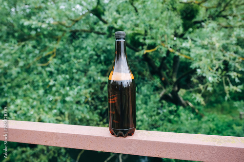 Dark brown bottle without the label standing on the iron fence in the green forest.