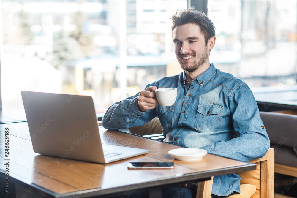 Portrait of handsome happy bearded young businessman in blue jeans shirt are sitting in cafe and have a break with cup of coffee and toothy smile are looking to the laptop. Portrait of handsome happy bearded young businessman in blue jeans shirt are sitting in cafe and have a break with cup of coffee and toothy smile are looking to the laptop.