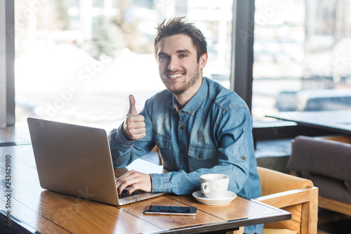 Everything alright! Portrait of handsome satisfied bearded young freelancer in blue jeans shirt are sitting in cafe and working on laptop with toothy smile and showing thumb up, looking at camera. © khosrork
