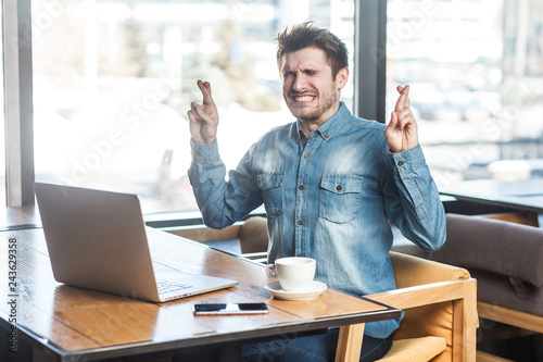 Praying! Side view portrait of wishful bearded young freelancer in blue jeans shirt are sitting in cafe and working on laptop, closed eyes and asking dream come true with clenched teeth. indoor