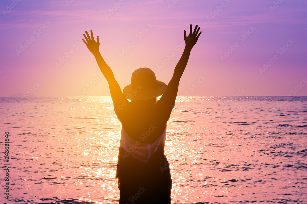 The silhouette of the happy woman holds arms up while on the seaside at the evening sunset,