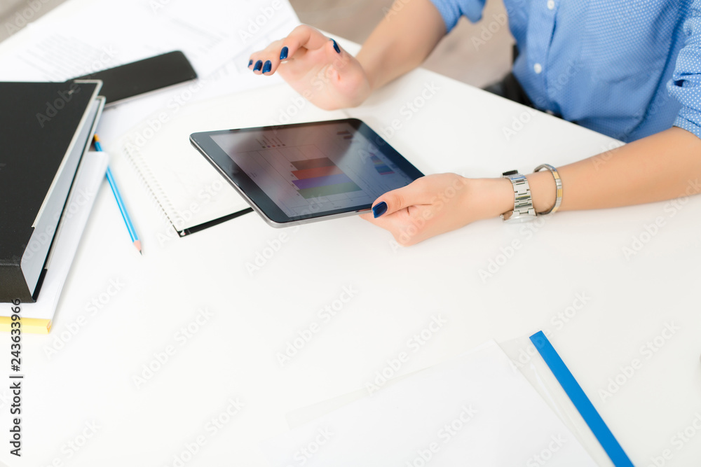 Girl's hands with blue manicure holding tablet at the white table in the office. She works with infographics. Girl's hands with blue manicure holding tablet at the white table in the office. She works with infographics.