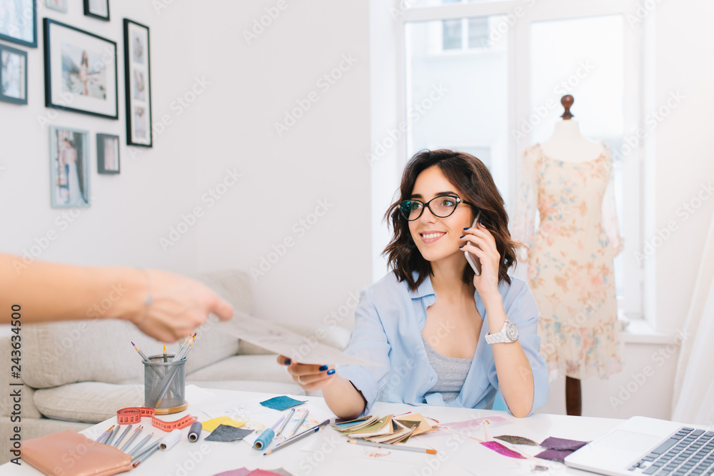 A cute  brunette girl in a blue shirt is siiting at the table in workshop.  She is speaking on phone and taking some sketch from client.
