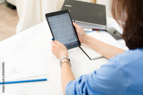 Girl is sitting at the white table  in office. She holds tablet and works with financial diagrams.