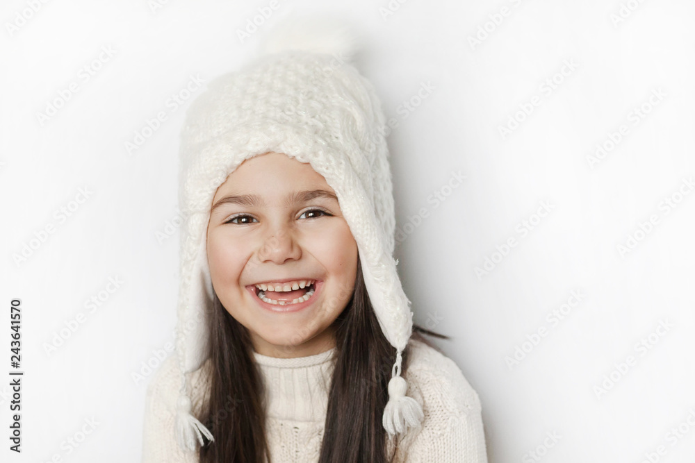 Happy cute child girl in a white winter hat and a white knitted sweater laughs on a white background. Real positive emotions. Smiling beautiful face. Happy cute child girl in a white winter hat and a white knitted sweater laughs on a white background. Real positive emotions. Smiling beautiful face.