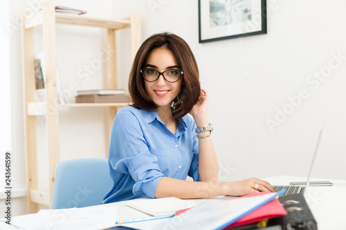 A pretty brunette girl in a blue shirt is sitting at the table in office. She works with laptop and  smiling cute to the camera.