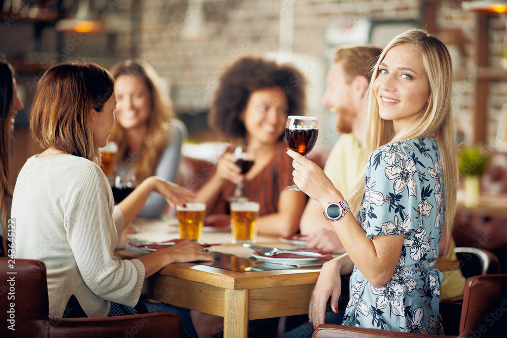 Woman looking at camera and holding glass of wine while sitting in restaurant. In background friends drinking and chatting.