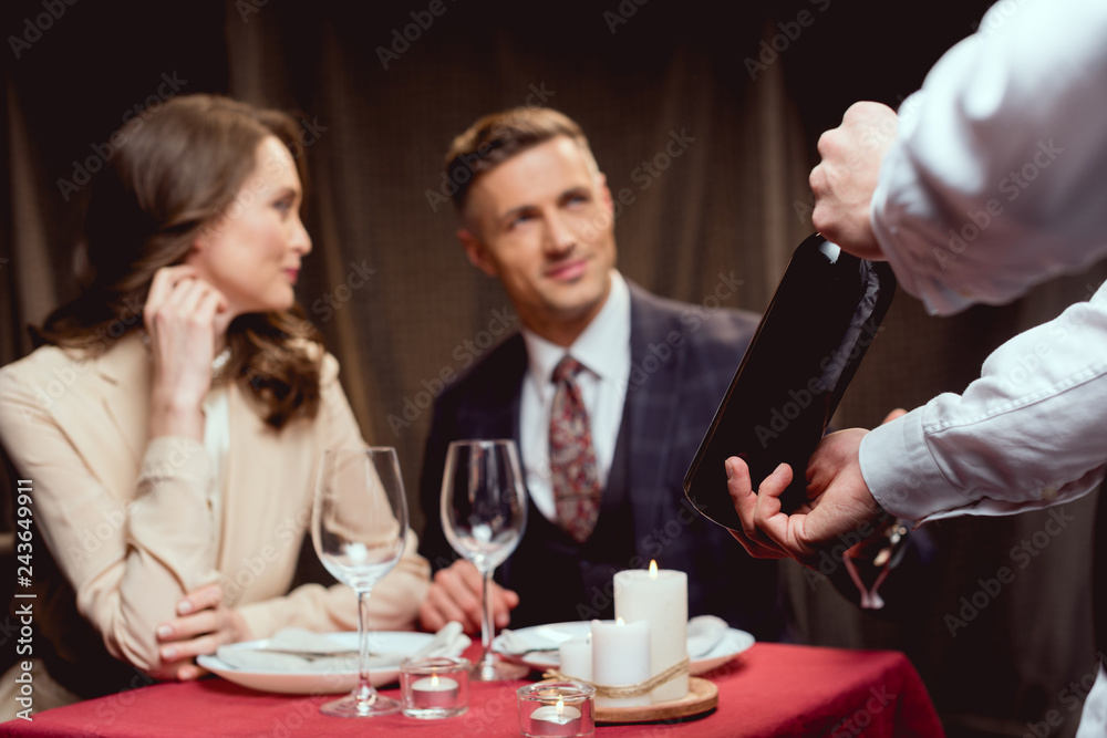 selective focus of waiter showing bottle of wine to beautiful couple during romantic date in restaurant selective focus of waiter showing bottle of wine to beautiful couple during romantic date in restaurant